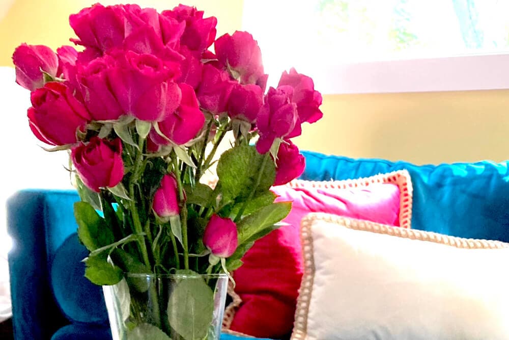 A bouquet of vibrant pink roses in a glass vase sits on a table with colorful cushions in the background.