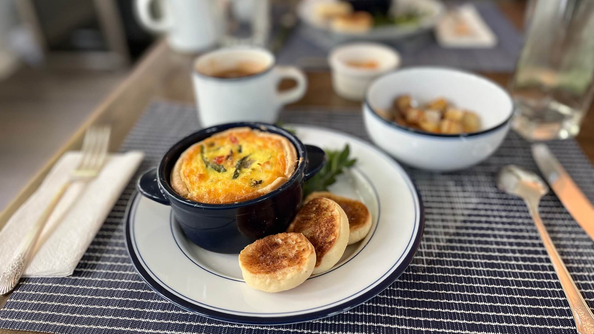 A plate featuring a mini vegetable quiche, toasted bread, and a bowl of roasted potatoes, alongside a cup of coffee.